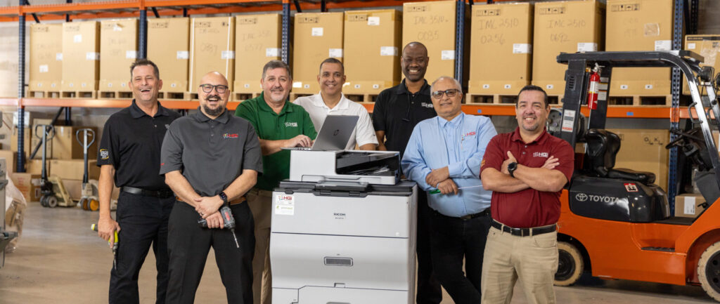 Our customers come first at HGi. Employees from HGI standing in the warehouse next to a copy machine.